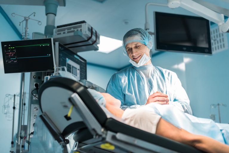 Un homme habillé avec des équipements de protections médicales sourie à une femmes couchée dans un salle d'opération