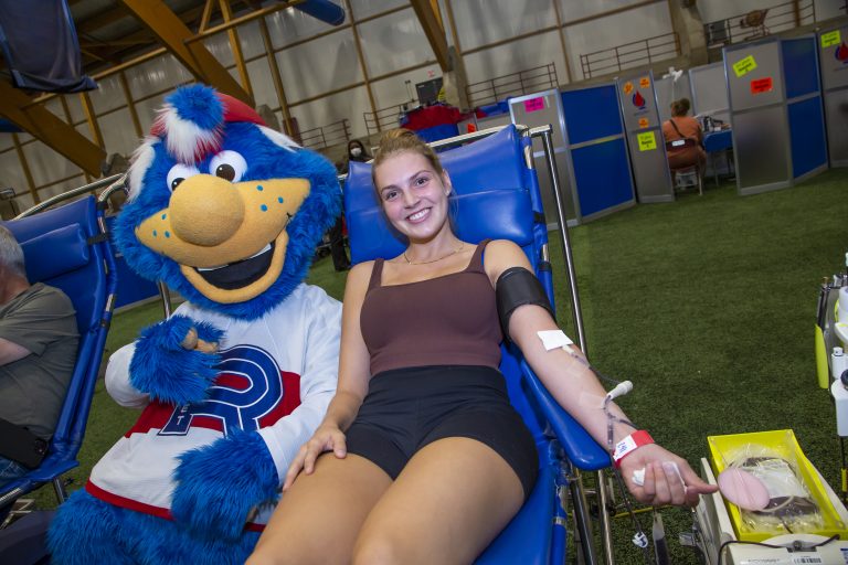 Jeune femme faisant un don de sang à la Collecte de sang des Lavallois. Elle est accompagné de Cosmo, la mascotte du Rocket de Laval.